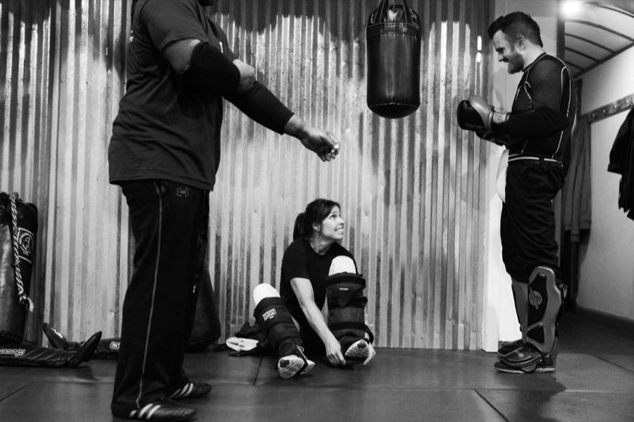A student sitting on the mat putting on shin guards while her training partners get ready by the heavy bag